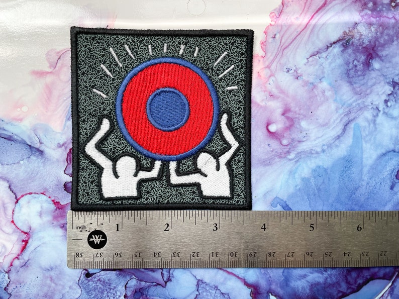 May include: A black and white embroidered patch featuring two figures holding up a red and blue circle with a smaller blue circle in the center. The circle has a white outline and is surrounded by white rays.