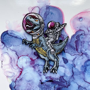May include: An embroidered patch featuring a blue dinosaur wearing a space helmet with a white astronaut riding on its back. The patch is set against a purple and blue watercolor background.