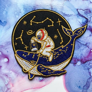May include: Embroidered patch featuring a white astronaut holding a lantern, riding on a blue whale with gold accents against a black background with gold stars and constellations.