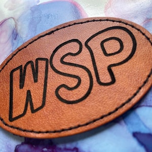 May include: A brown leather patch with black stitching around the edges. The patch has the letters "WSP" in black outlined letters.