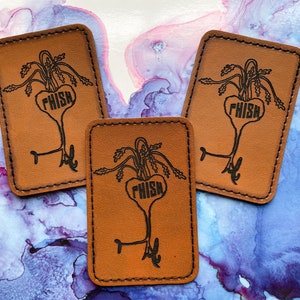 May include: Three brown leather patches with a black outline and the word "PHISH" in a heart shape. The patches feature a stylized image of a plant with a heart-shaped base.