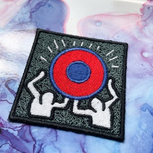 May include: Embroidered patch with two white figures holding a red and blue circle with a white outline. The background is black with a green leafy pattern.