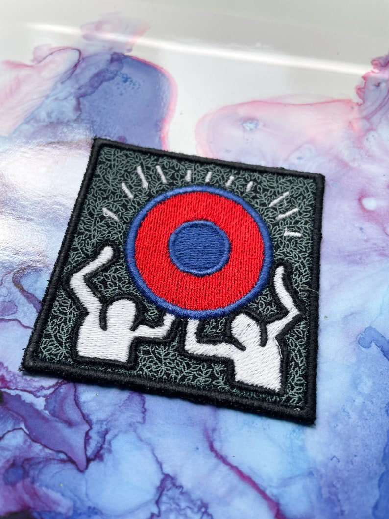 May include: Embroidered patch with two white figures holding a red and blue circle with a white outline. The background is black with a green leafy pattern.