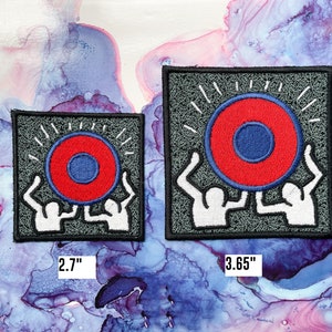 May include: Two white figures holding up a red and blue circle with a white outline. The circle has a smaller blue circle in the center. The figures and circle are embroidered on a gray fabric patch. The patch is 2.7 inches wide and 3.65 inches tall.