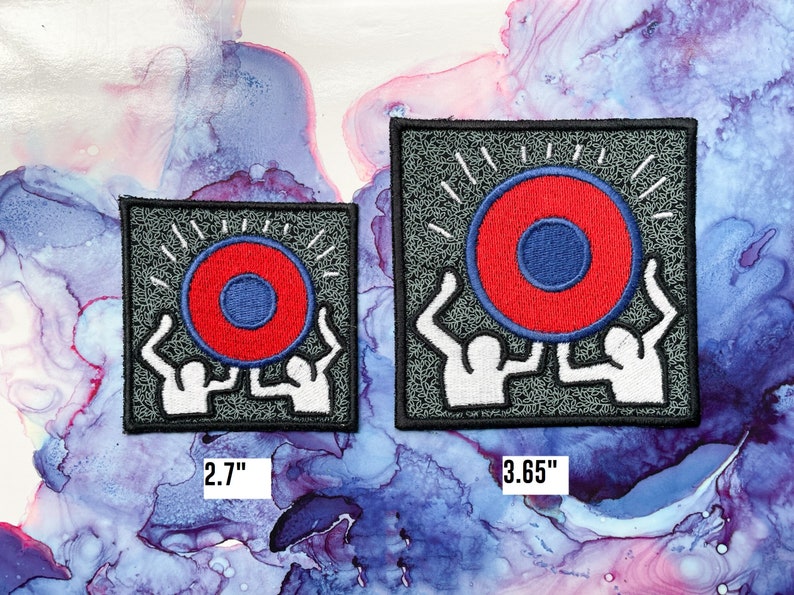May include: Two white figures holding up a red and blue circle with a white outline. The circle has a smaller blue circle in the center. The figures and circle are embroidered on a gray fabric patch. The patch is 2.7 inches wide and 3.65 inches tall.