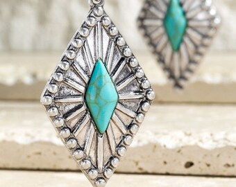 Puede incluir: Pendientes plateados con una piedra de color turquesa en el centro. Los pendientes tienen forma de diamante con un diseño de rayos de sol y un borde de cuentas. La parte superior del pendiente tiene un diseño floral.
