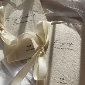 May include: Two ivory wedding cards tied with cream-colored satin ribbons. The cards read "To my husband" and "To my wife" with the date 08.10.2025 and initials "C&B". The cards are on a white tulle background.