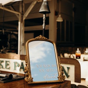 Puede incluir: Espejo rectangular con marco dorado y detalles ornamentados. El espejo refleja un cielo azul con nubes y el texto "YOU LOOK LIKE YOU NEED A drink". El espejo está sobre una superficie de madera.