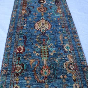 Puede incluir: Una alfombra azul y roja con un diseño floral. La alfombra tiene un borde con flecos y está hecha de lana.