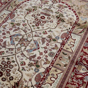 May include: A detailed beige rug with a central floral pattern and a deep red border. The rug displays intricate patterns of flowers, birds, and geometric shapes in shades of red, green, and blue. The rug's design is symmetrical and detailed.
