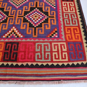 May include: A vibrant, geometric patterned rug. The rug features a central blue field with orange, black, and red geometric shapes. The border is red with repeating square patterns. The rug has a fringed edge. The rug's measurements are in centimetres.