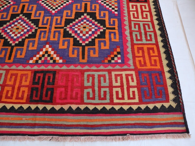 May include: A vibrant, geometric patterned rug. The rug features a central blue field with orange, black, and red geometric shapes. The border is red with repeating square patterns. The rug has a fringed edge. The rug's measurements are in centimetres.