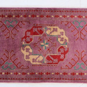 May include: A vintage rug with a pink background and intricate patterns in red, cream, and green. The rug has a geometric design with floral motifs.