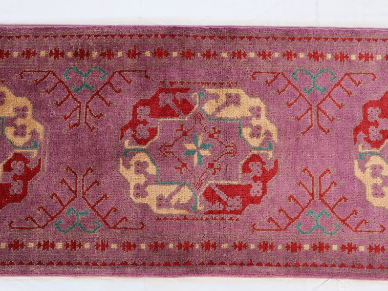 May include: A vintage rug with a pink background and intricate patterns in red, cream, and green. The rug has a geometric design with floral motifs.
