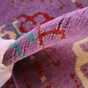May include: Close-up of a purple, red, and cream colored rug with a hand holding the edge of the rug. The rug has a traditional Persian design.