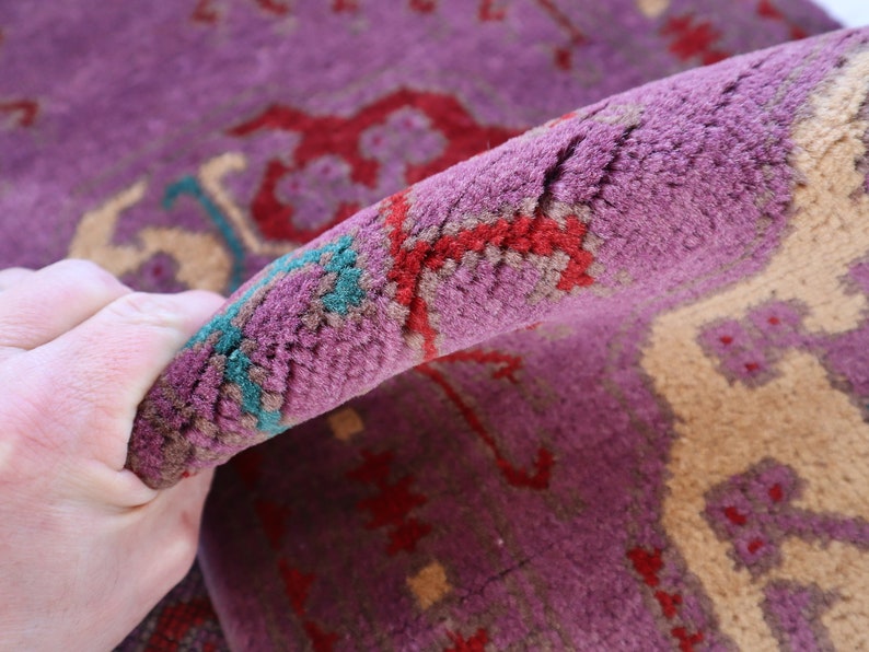 May include: Close-up of a purple, red, and cream colored rug with a hand holding the edge of the rug. The rug has a traditional Persian design.