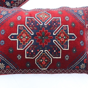 May include: Rectangular decorative pillow with a red background and a central geometric design in navy blue, white, and green. The pillow features a detailed, patterned border and a symmetrical floral motif.