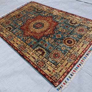May include: Rectangular area rug with a central medallion design in red and gold, set against a teal background. The rug features intricate patterns and a cream-colored border, with colorful fringe along the edges. A decorative floor covering.