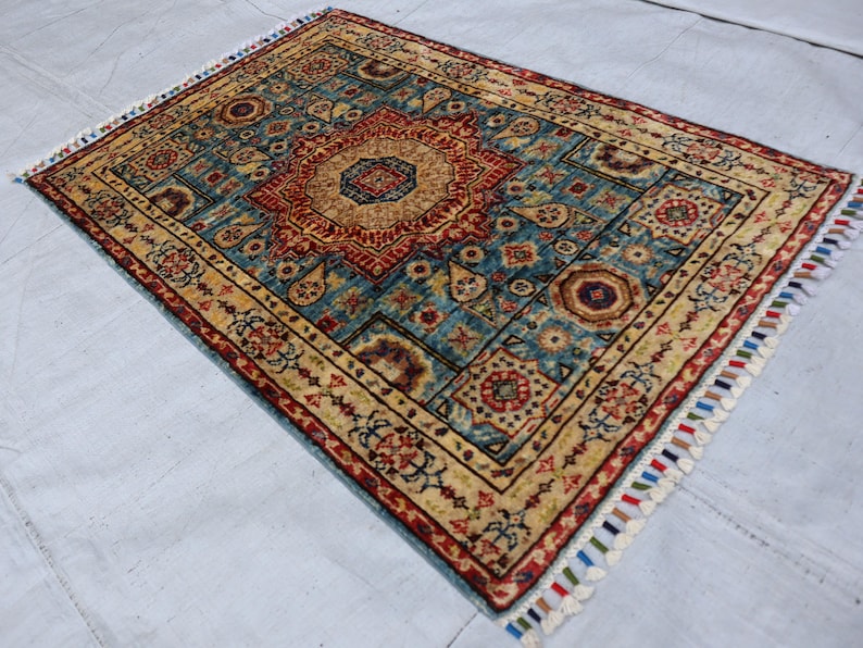 May include: Rectangular area rug with a central medallion design in red and gold, set against a teal background. The rug features intricate patterns and a cream-colored border, with colorful fringe along the edges. A decorative floor covering.