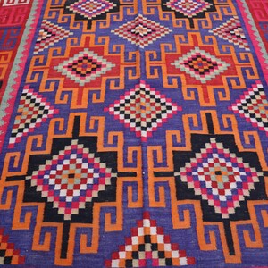 May include: A colourful, hand-woven rug with a geometric design. The rug has a central purple field with orange, black, red, and white geometric shapes. The border is red and black with repeating patterns. This rug is ideal for adding colour and texture to a room.
