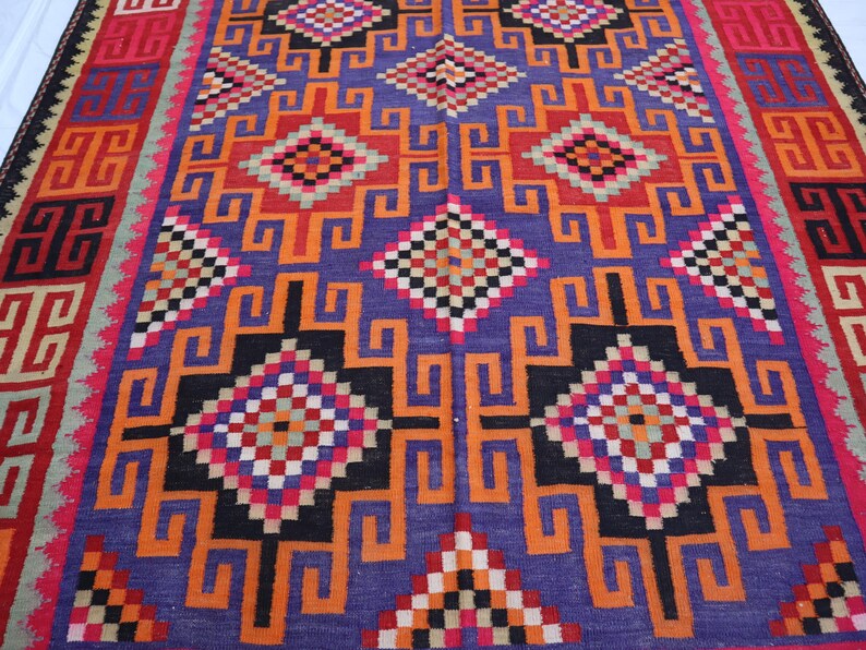 May include: A colourful, hand-woven rug with a geometric design. The rug has a central purple field with orange, black, red, and white geometric shapes. The border is red and black with repeating patterns. This rug is ideal for adding colour and texture to a room.