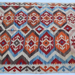 May include: Hand-woven rug with a geometric pattern. The rug features repeating diamond and hexagonal shapes in shades of red, orange, blue, brown, and beige. The rug has a fringed edge and is suitable for home decor.