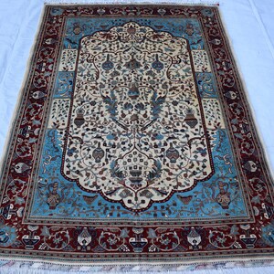 May include: An intricately patterned rectangular rug. The central cream field displays a floral and vase design in blue, brown, and green. The border is a deep red with detailed patterns, and the rug has a fringed edge.