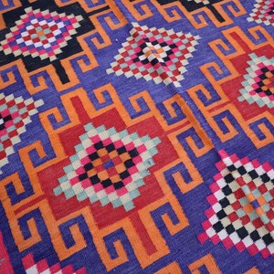 May include: Close-up of a colourful woven rug with a geometric pattern. The rug features a repeating design of squares and stylized shapes in shades of blue, orange, red, black, and pink.