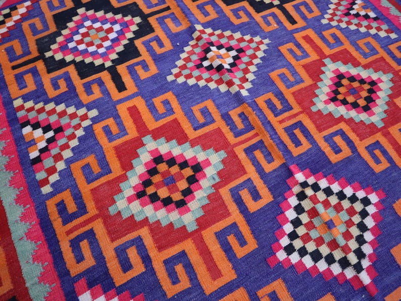 May include: Close-up of a colourful woven rug with a geometric pattern. The rug features a repeating design of squares and stylized shapes in shades of blue, orange, red, black, and pink.