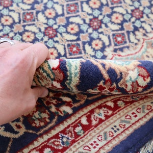 Puede incluir: Primer plano de una alfombra azul y roja con estampado que se est&aacute; doblando con una mano. La alfombra tiene un dise&ntilde;o floral detallado.