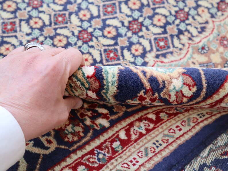 Puede incluir: Primer plano de una alfombra azul y roja con estampado que se est&aacute; doblando con una mano. La alfombra tiene un dise&ntilde;o floral detallado.