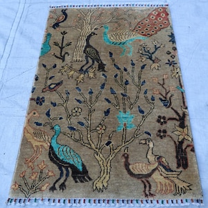 May include: A rectangular, beige rug with a tree and bird design. The rug features various birds in teal, black, and brown, with a tree in the center. The rug has a fringe border with red, white, and blue accents. The rug is likely used as a decorative floor covering.