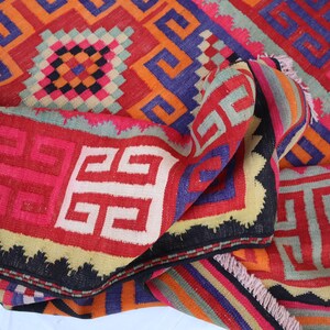 May include: A folded, colourful rug with geometric patterns in red, orange, blue, and black. The rug features repeating square and swirled designs, with a fringed edge.