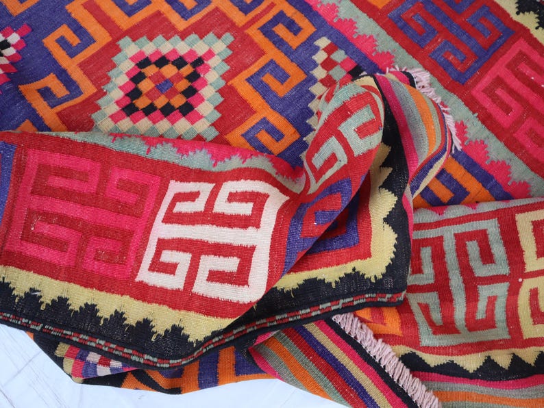 May include: A folded, colourful rug with geometric patterns in red, orange, blue, and black. The rug features repeating square and swirled designs, with a fringed edge.