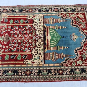 May include: A traditional prayer rug with a red and blue design. The rug features a mosque with a green dome and intricate floral patterns.