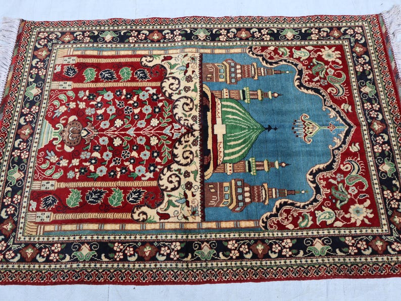 May include: A traditional prayer rug with a red and blue design. The rug features a mosque with a green dome and intricate floral patterns.