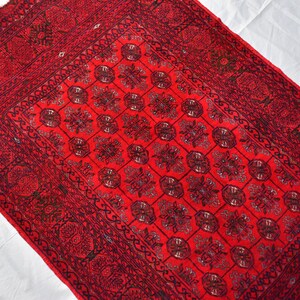May include: A rectangular, red rug with an intricate, repeating floral pattern. The rug features a dark border with detailed designs and a central field of repeating floral medallions. The rug is likely made of woven material.