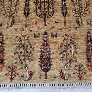 Könnte beinhalten: Beigefarbener Teppich mit einem sich wiederholenden Muster aus stilisierten Bäumen und Tieren. Die Bäume haben rote, blaue und braune Blätter mit kleinen Tiermotiven. Der Teppich hat einen Fransenrand.