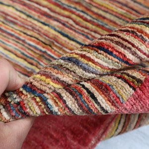 May include: Close-up of a multi-colored striped rug with a hand holding the edge of the rug. The rug has a variety of colors including red, orange, yellow, green, blue, and purple.