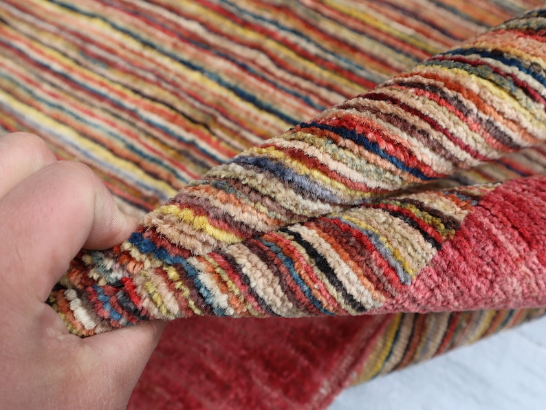 May include: Close-up of a multi-colored striped rug with a hand holding the edge of the rug. The rug has a variety of colors including red, orange, yellow, green, blue, and purple.
