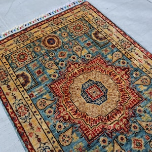 May include: A rectangular rug with an intricate pattern. The rug features a central medallion in shades of red, tan, and blue, surrounded by a border of tan and blue geometric designs. The rug has a fringe on one end.