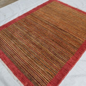 May include: A rectangular rug with a red border and a striped pattern in shades of brown, beige, and black.