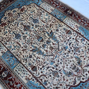 May include: An intricately designed rug with a cream-colored central field, adorned with floral and vase motifs in shades of blue, brown, and beige. The border features a deep red and light blue pattern, creating a classic, elegant aesthetic.