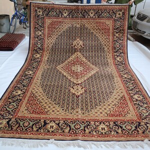 Puede incluir: Una alfombra grande con un patr&oacute;n intrincado, fondo azul oscuro, acentos beige y rojo y un dise&ntilde;o central en forma de diamante. La alfombra se est&aacute; desenrollando sobre una superficie blanca.