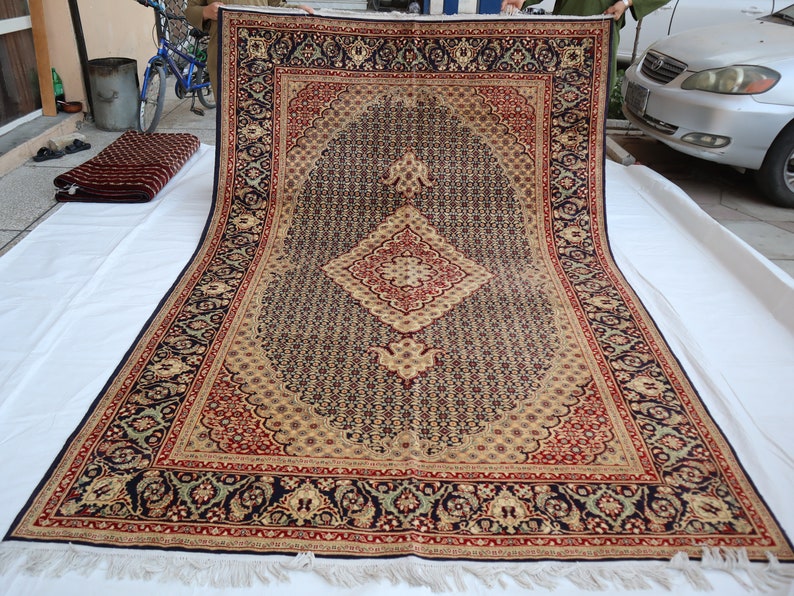 Puede incluir: Una alfombra grande con un patr&oacute;n intrincado, fondo azul oscuro, acentos beige y rojo y un dise&ntilde;o central en forma de diamante. La alfombra se est&aacute; desenrollando sobre una superficie blanca.