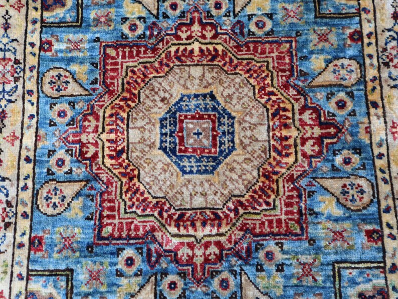 May include: Close-up of a detailed, patterned rug with a central medallion design. The rug features a vibrant color palette of blue, red, beige, and gold. The intricate patterns include geometric shapes and floral motifs, creating a rich, textured appearance.