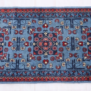Op de afbeelding: Een blauw loperkleed met een herhalend patroon van bloemen en geometrische ontwerpen in rood, marineblauw en crème. Het kleed heeft een rand met een herhalend patroon in rood en marineblauw. Het kleed is waarschijnlijk gemaakt van wol of een vergelijkbaar materiaal.