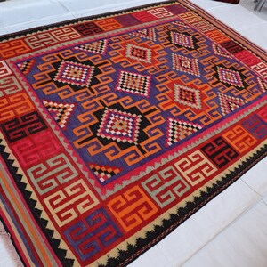 its 7x10 ft rug, A vibrant, rectangular rug with a geometric pattern. The rug features a central field of purple with orange and black diamond shapes. The border is red with repeating geometric designs. The rug is made of woven wool