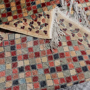 May include: A beige rug with a checkered pattern in shades of red, blue, and green. The rug has a fringe border.
