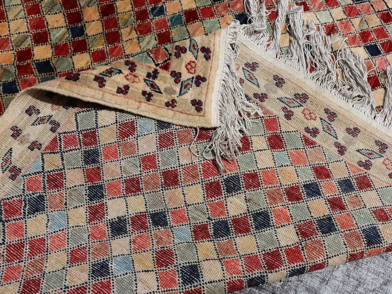 May include: A beige rug with a checkered pattern in shades of red, blue, and green. The rug has a fringe border.
